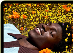 MacBook Neo, couleur jaune agrume, écran affichant une personne souriante allongée dans un champ de fleurs ensoleillé