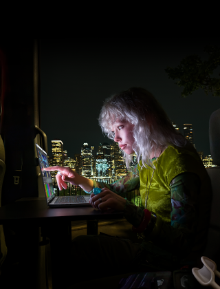 Une personne est assise sur la banquette arrière d’une camionnette, la porte passager ouverte laisse voir la ligne d’horizon d’une ville la nuit, un MacBook Pro avec Écran PROLED XDR est posé sur une table de travail, la personne, éclairée par l’écran, utilise la fonctionnalité tactile (Touch) pour monter une vidéo dans DaVinci Resolve Studio