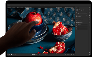 MacBook Pro avec Écran PROLED XDR affichant une image de nature morte de grenades avec des céramiques et des textiles bleus, une main se tend vers l’écran pour interagir avec l’image